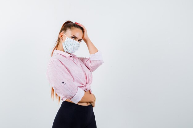 Portrait of young female holding hand on head in shirt, pants, medical mask and looking confident front view