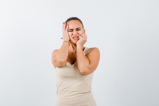 Portrait of young female holding hands on cheeks in beige tank top and looking displeased front view