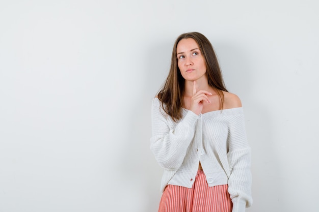 Portrait of young female keeping finger under chin in cardigan isolated