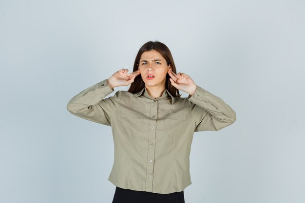 Portrait of young female keeping fingers behind ears while frowning in shirt, skirt and looking confused front view