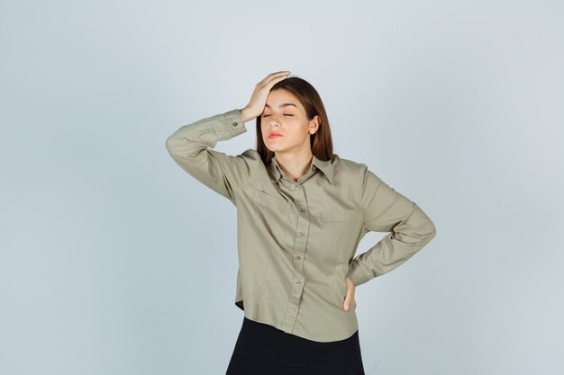 Portrait of young female keeping hand on head in shirt, skirt and looking tired front view