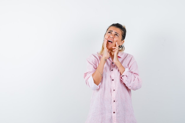 Portrait of young female keeping hands on cheeks in pink shirt and looking worried front view