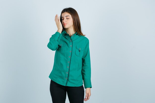Portrait of young female keeping palm in front of face in green shirt, pants and looking delighted front view