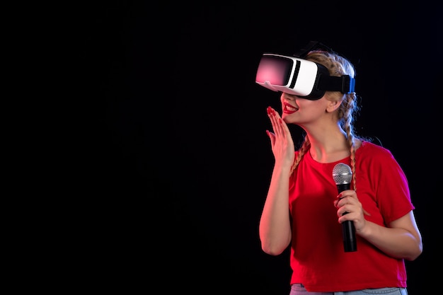 Portrait of young female playing vr and singing with mic on a dark wall