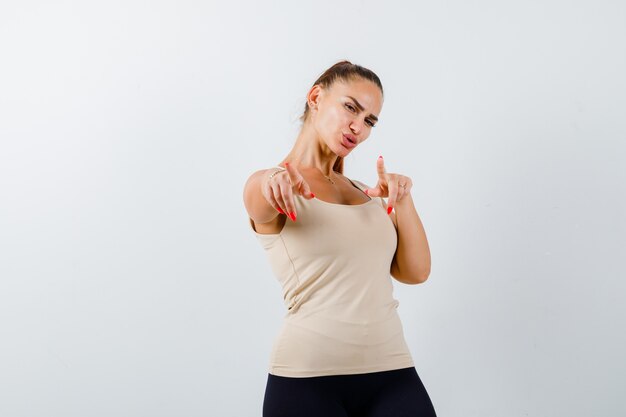 Portrait of young female pointing at camera in beige tank top and looking confident front view