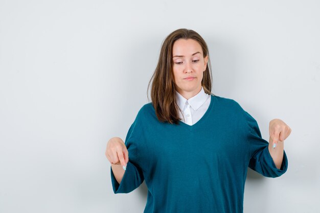 Portrait of young female pointing down in sweater over shirt and looking displeased front view