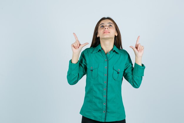Портрет молодой женщины, указывая вверх в зеленой рубашке и смотрящий обнадеживающий вид спереди