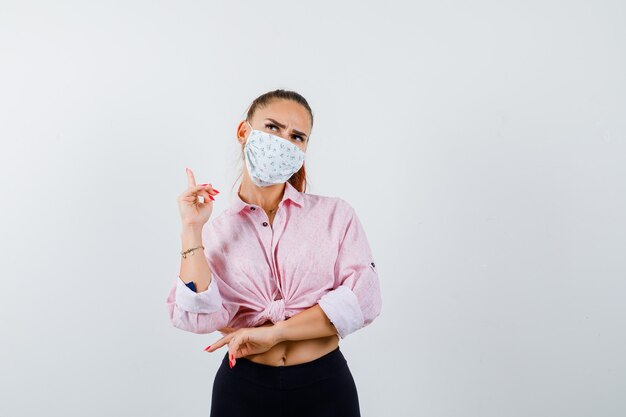 Portrait of young female pointing up in shirt, pants, medical mask and looking pensive front view