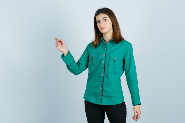 Portrait of young female pointing at upper left corner in green shirt, pants and looking confused front view