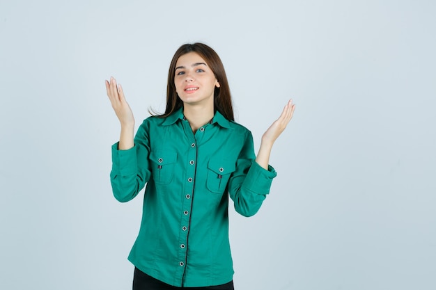 Портрет молодой женщины, поднимающей руки, улыбаясь в зеленой рубашке и глядя обнадеживающий вид спереди