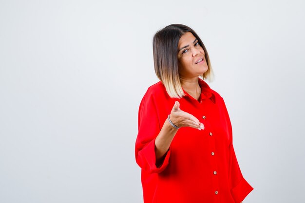 Портрет молодой женщины, протягивающей руку для приветствия в красной негабаритной рубашке и уверенно выглядящей, вид спереди
