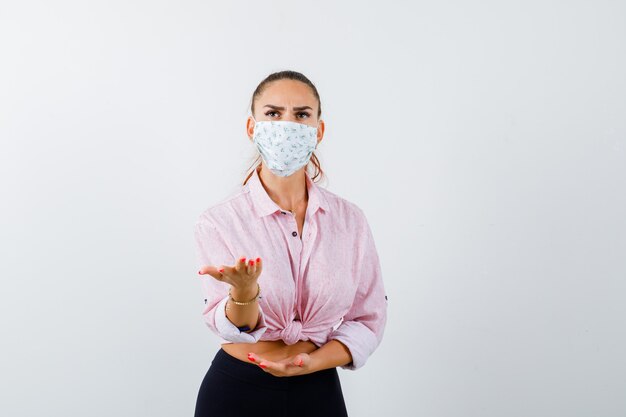 Portrait of young female stretching hand in puzzled gesture in shirt, pants, mask and looking angry front view