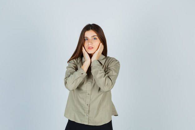 Portrait of young female touching her cheeks with hands in shirt, skirt and looking graceful front view