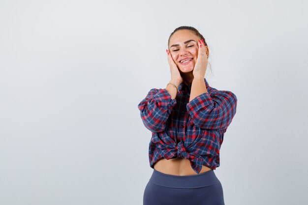 Portrait of young female with hands on cheeks in checkered shirt, pants and looking happy front view