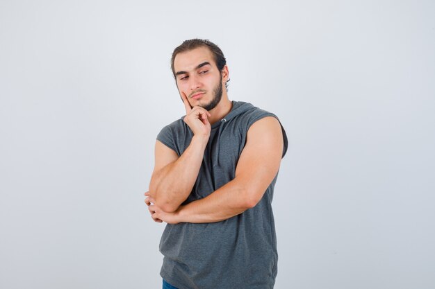 Portrait of young fit male keeping finger on cheek in sleeveless hoodie  and looking pensive front view