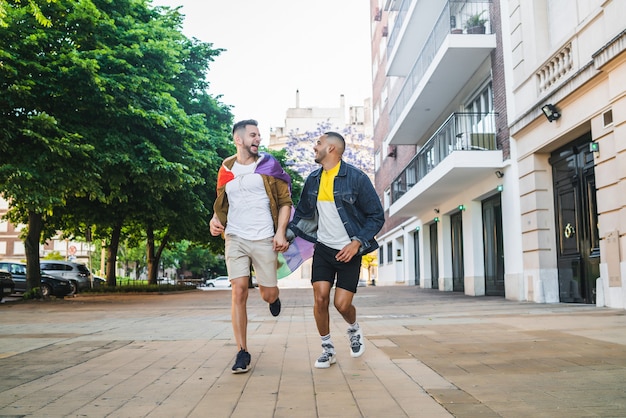 手をつないで、通りで虹色の旗と一緒に走っている若い同性愛者のカップルの肖像画。 LGBTと愛の概念。