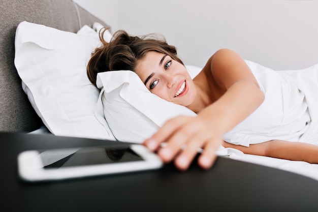 Портрет молодой девушки на кровати в современной квартире утром. Она берет телефон на столе и улыбается.