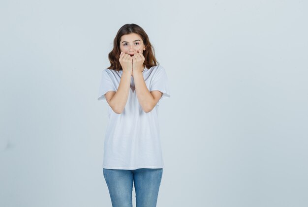 Portrait of young girl biting fists emotionally in white t-shirt and looking scared front view