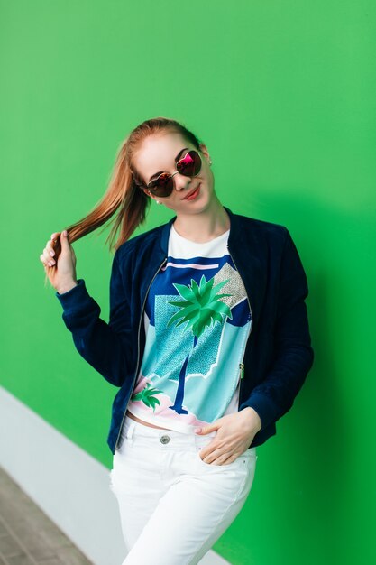 A portrait of a young girl in blue jacket outdoor near green wall with white line down. The girl wears sunglasses, holds hair tail in hand and looking to the camera.