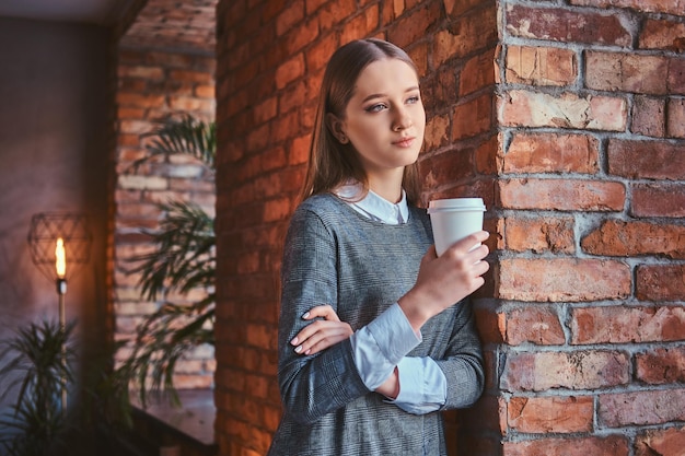 Портрет молодой девушки, одетой в элегантное серое платье, прислоненной к кирпичной стене, держит чашку кофе на вынос и смотрит в сторону.
