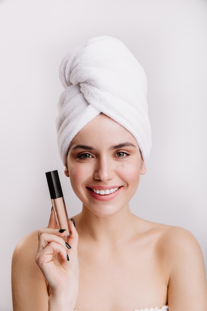 Portrait of young girl in great mood on white wall. Woman with towel on her head posing with concealer.