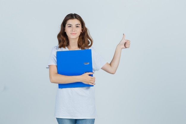 Portrait of young girl holding folder, showing thumb up in white t-shirt and looking jolly front view