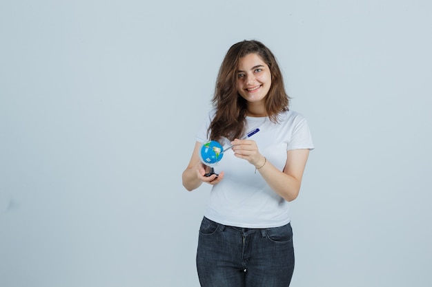 Portrait of young girl holding pen on globe in t-shirt, jeans and looking happy front view