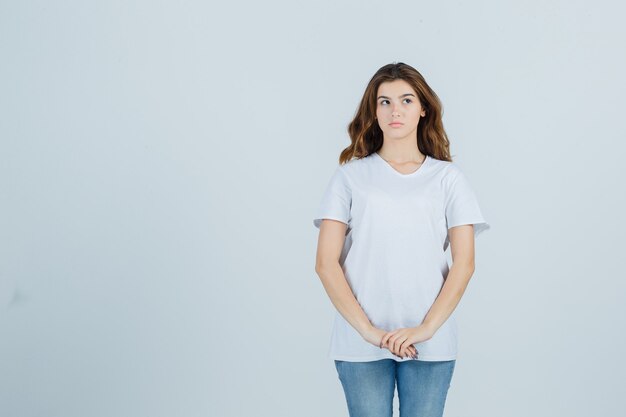 Портрет молодой девушки, держащей руки перед собой в белой футболке и задумчивой смотрящей спереди