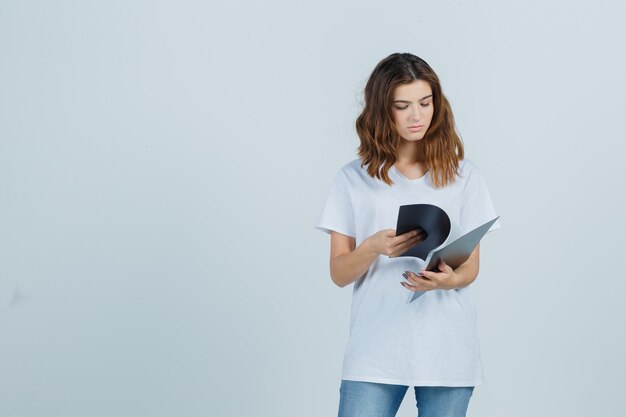 Портрет молодой девушки просматривает заметки в папке в белой футболке и смотрит сосредоточенный вид спереди
