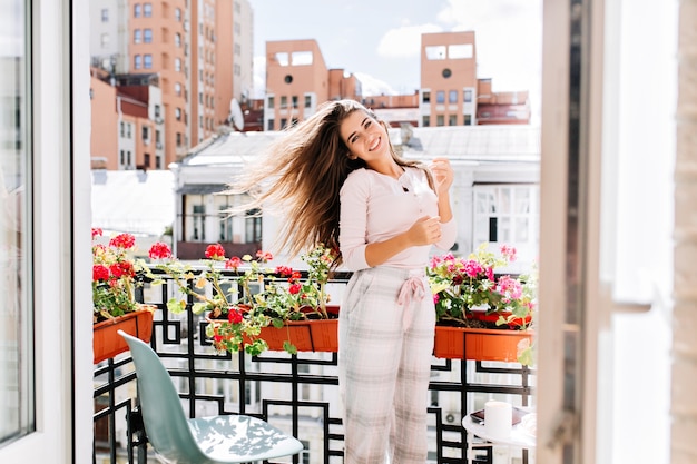 Portrait young girl in pajama moving on balcony in city in sunny morning. Her long hair flying on wind, she smiling .