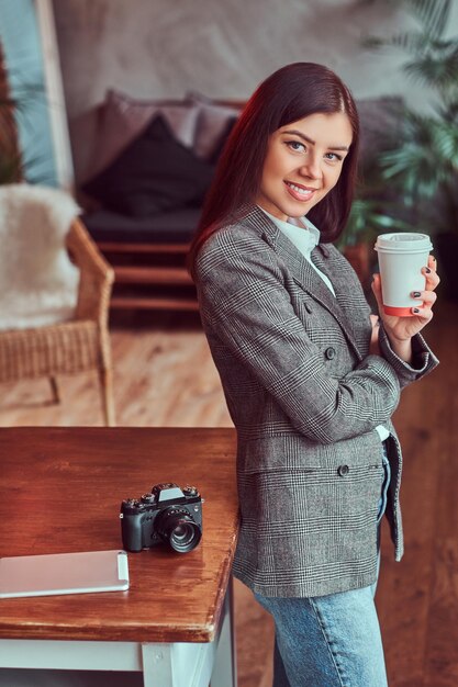 カメラを見ながら、ロフトインテリアの部屋のテーブルに寄りかかってテイクアウトコーヒーのカップを保持している灰色のエレガントなジャケットに身を包んだ若い女の子の写真家の肖像画。