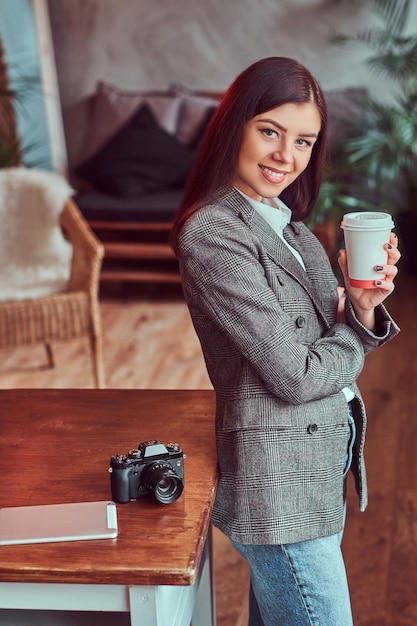 Портрет молодой девушки-фотографа, одетой в серый элегантный жакет, держащей чашку кофе на вынос, опираясь на стол в комнате с лофтовым интерьером, глядя в камеру.