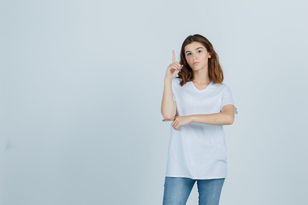 Портрет молодой девушки, указывающей вверх в белой футболке и выглядящей разумно, вид спереди