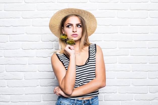 Portrait young girl posing in straw hat enjoying summer while standing on white brick wall