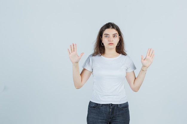 Портрет молодой девушки, поднимающей руки в жесте капитуляции в футболке, джинсах и серьезного вида спереди