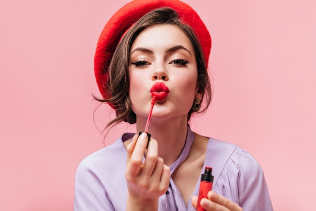 Portrait of young girl in red beret painting her lips with bright lipstick on pink background.