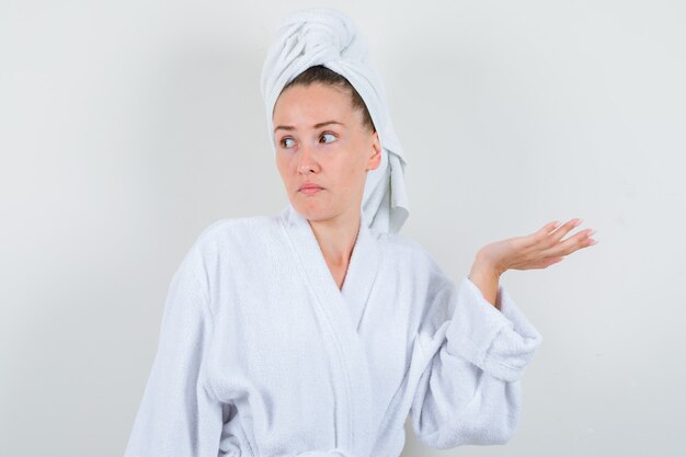 Portrait of young girl showing helpless gesture in white bathrobe, towel and looking confused front view