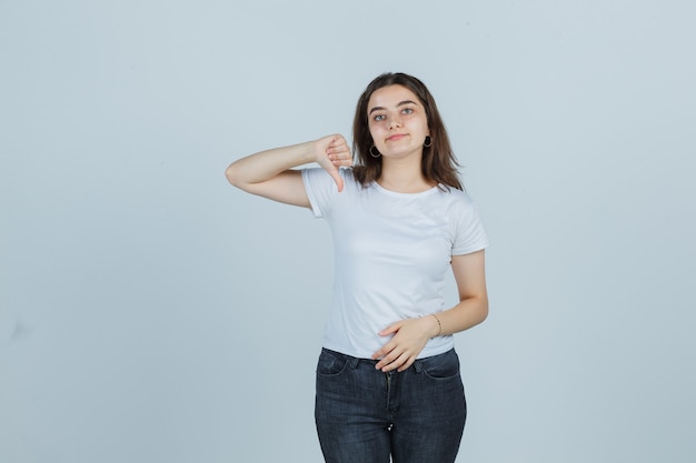 Портрет молодой девушки, показывающей большой палец вниз в футболке, джинсах и уверенно выглядящей вид спереди