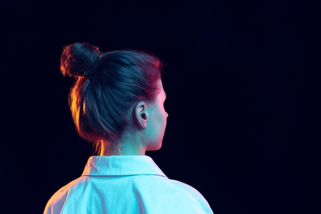 Ritratto di giovane ragazza in camicia bianca in posa isolata su sfondo blu scuro in luce al neon vista posteriore