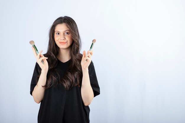 Portrait of young girl with cosmetic brushes looking at camera on white.