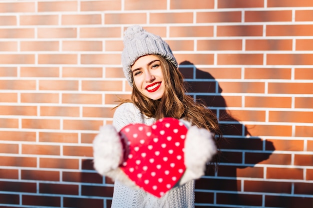 Бесплатное фото Портрет молодой девушки с длинными волосами в вязаной шапке, теплом свитере на стене снаружи. она протягивает красное сердце в перчатках.