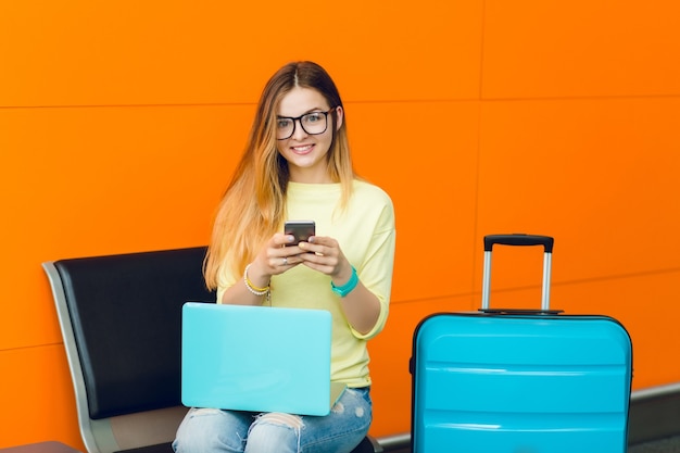 Portrait of young girl in yellow sweater sitting on chair on orange background. She has blue laptop on knees and blue suitcase near. She is smiling to the camera.