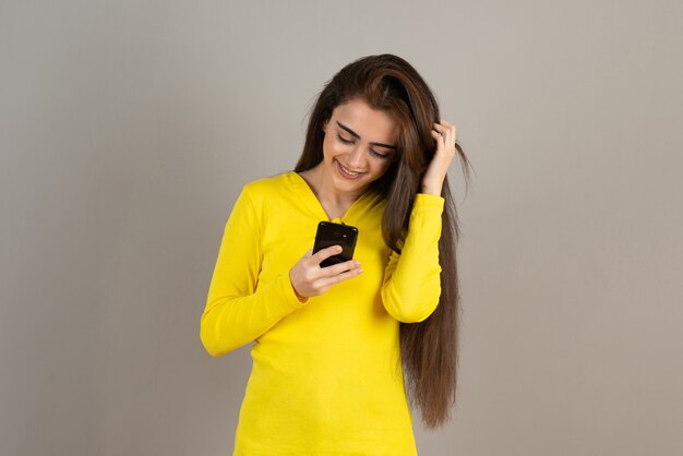 Portrait of young girl in yellow top holding cellphone on gray wall.