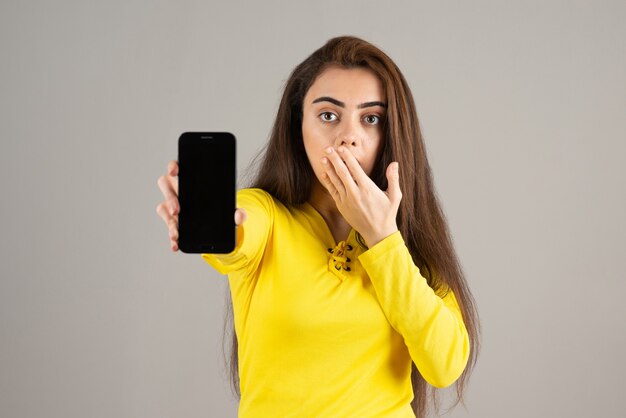 Portrait of young girl in yellow top posing with cellphone on gray wall.