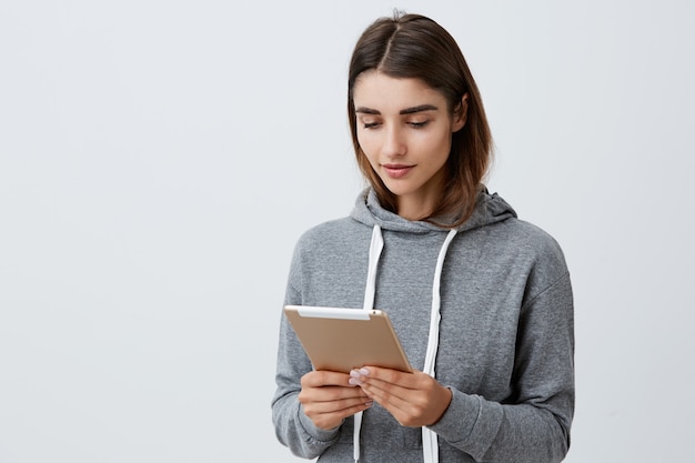 Portrait of young good-looking caucasian student girl with dark long hair in casual gray hoodie holding digital tablet in hands, reading tasks for university, getting ready for exam. Urban lifestyle.