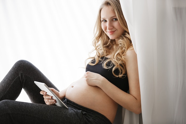 Free photo portrait of young good-looking pregnant woman with blond hair in home clothes sitting on window in light bedroom