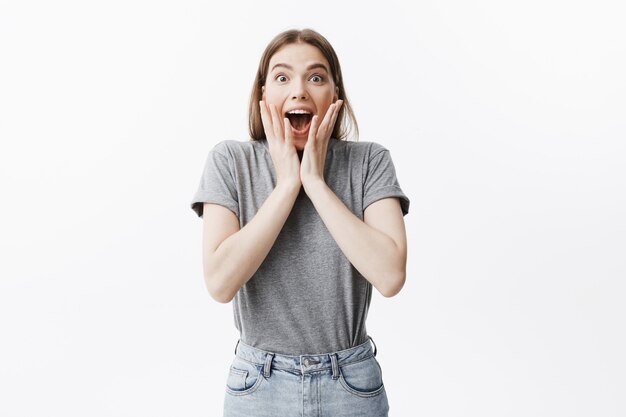 Portrait of young good looking student girl with dark hair in fashionable clothes squeezing face with hands in excitement with opened mouth seeing pop star on street