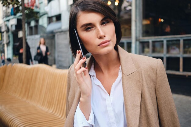 Portrait of young gorgeous businesswoman talking on cellphone thoughtfully looking in camera in cafe on street