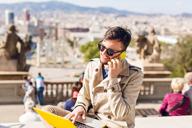 Portrait of young guy in beige coat sitting on hight on city background. He wears beige coat, working on yellow laptop, speaking on phone.