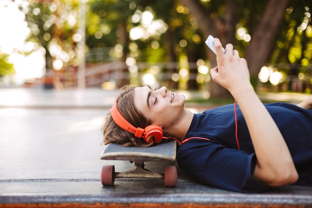 スケートパークで時間を過ごして携帯電話を楽しく使用しながらスケートボードに横たわっているオレンジ色のヘッドフォンで若い男の肖像画